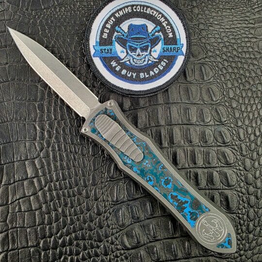 Hawk Deadlock Model C open on its show side with an M390 double-edge dagger and marbled blue Fat Carbon inlay beside the We Buy Knife Collections patch