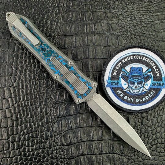 Hawk Deadlock Model C open on its clip side, with the Titanium pocket clip, fluted panel and blue Fat Carbon inlay, beside the We Buy Knife Collections patch