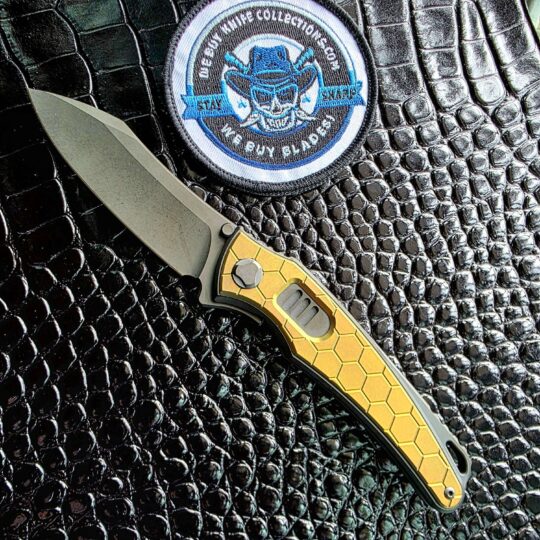 Hawk Talon Auto Blade Show West 2026 exclusive open on the show side with gold anodized honeycomb titanium inlays, We Buy Knife Collections patch in frame