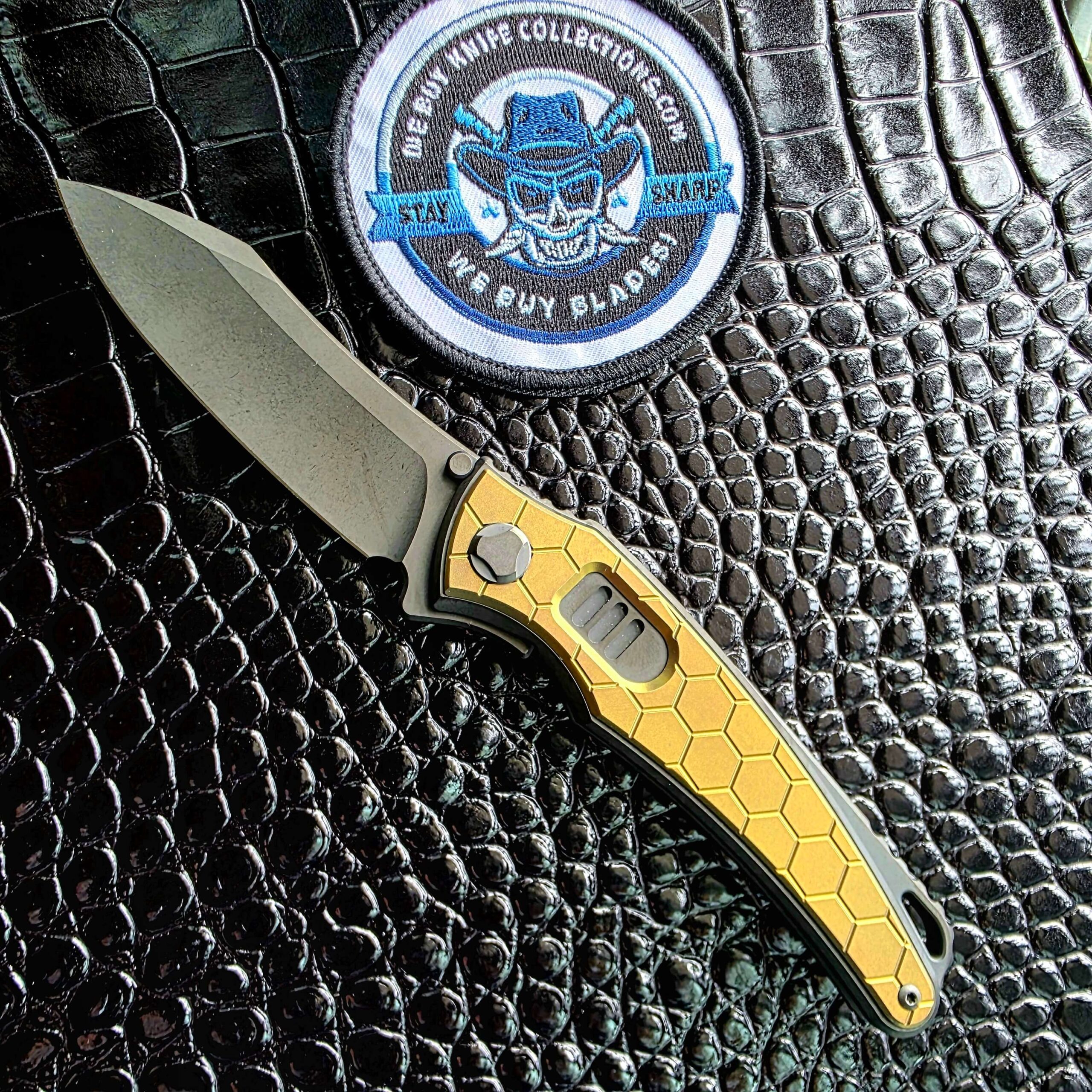 Hawk Talon Auto Blade Show West 2026 exclusive open on the show side with gold anodized honeycomb titanium inlays, We Buy Knife Collections patch in frame