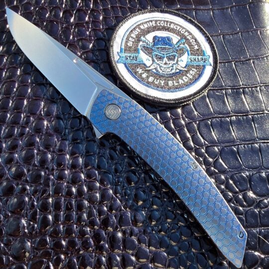 Hog House Honeycomb Model T Gen 2 serial 446 open on the show side in blue anodized titanium, We Buy Knife Collections patch in frame