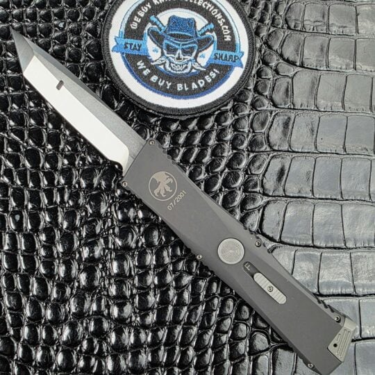 Microtech Exodus single action OTF open on its show side beside the We Buy Knife Collections patch