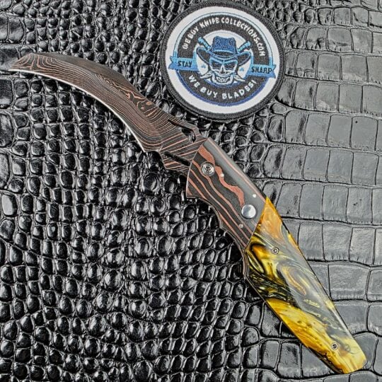 Reese Weiland Harpy open on its show side with a Brad Vice Copper Damascus hawkbill and Honey Hive Acrylic scale beside the We Buy Knife Collections patch