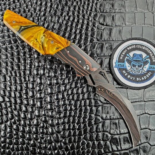 Reese Weiland Harpy open on its reverse side, with the Copper Damascus bolster and Honey Hive Acrylic, beside the We Buy Knife Collections patch